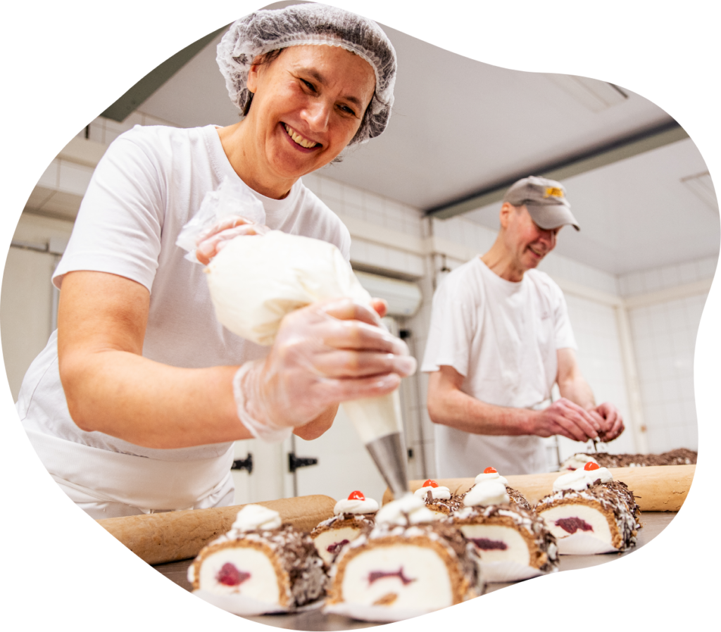 Eine Konditorin(ca. 48 Jahre alt) und ein Konditor (ca. 58 Jahre Alt) genieren freudig mit Konzentration, Kuchenstücke in Arbeitskleidung in der Backstube.