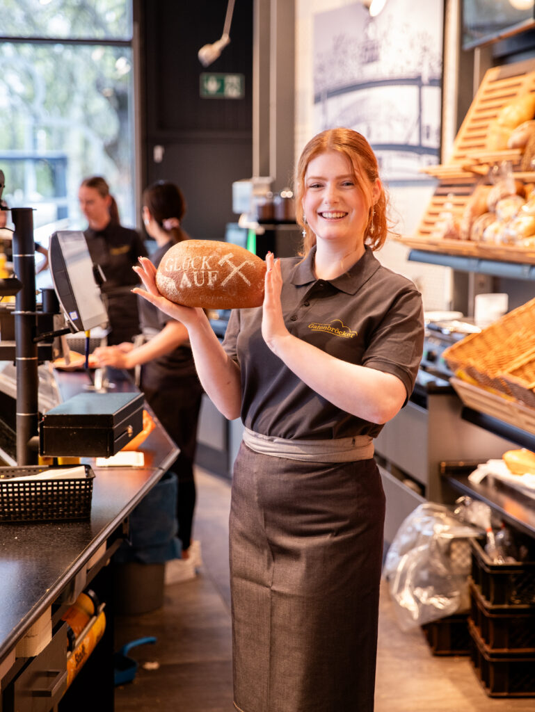 Freundliche Fachverkäuferin-Auszubildende im Alter von ca. 25 Jahren die in Arbeitskleidung hinter der Theke einer Filiale steht und ein gluck-Auf-Brot_Laib hält.