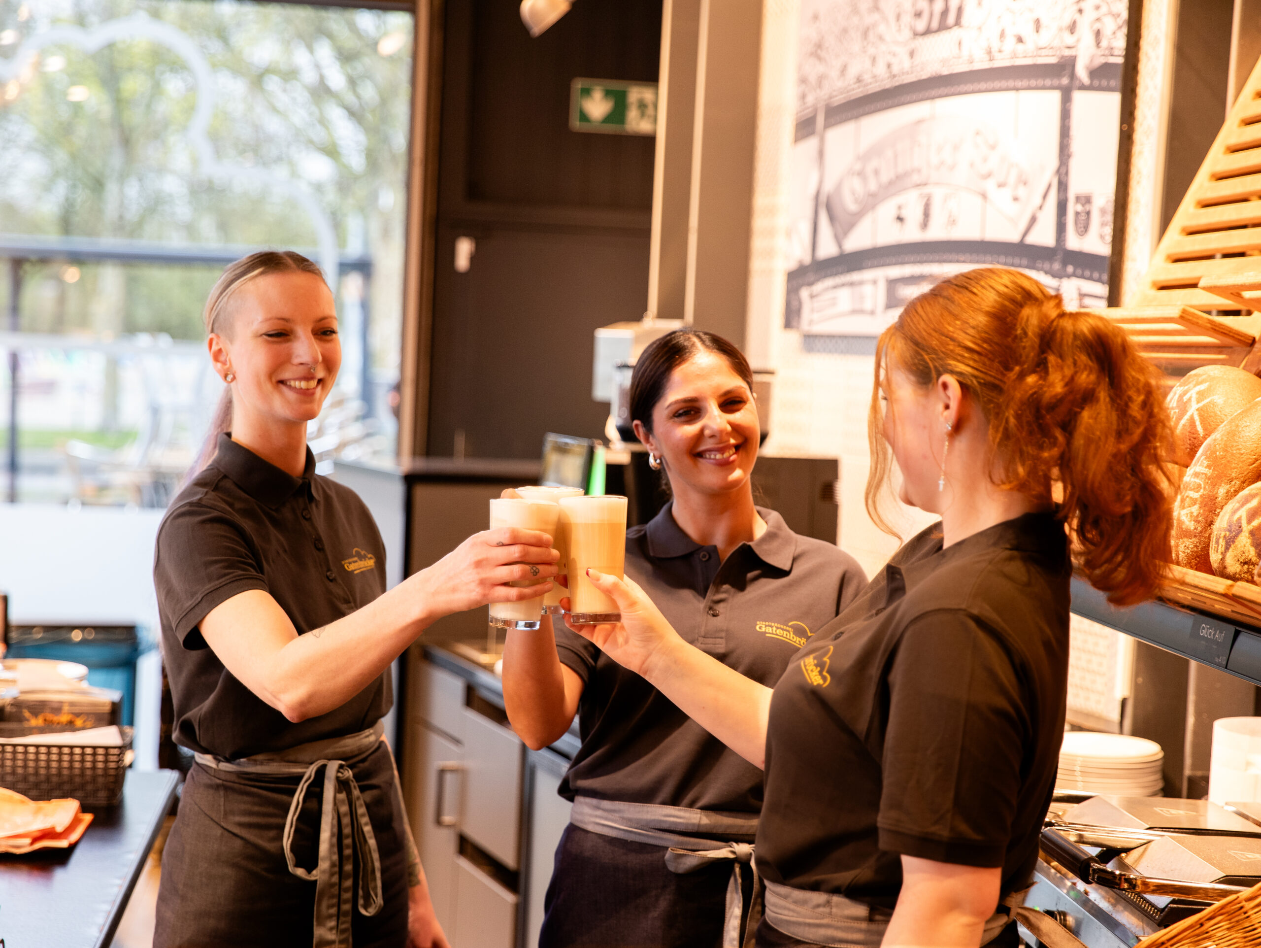 Drei Fachverkäuferinnen von Gatenbröcker ,in dem Alter von 25-35 Jahre, mit Firmen Polo & Taillenschütze, die lächeln und mit Cappuccino in einer Gatenbröcker Filiale anstoßen.